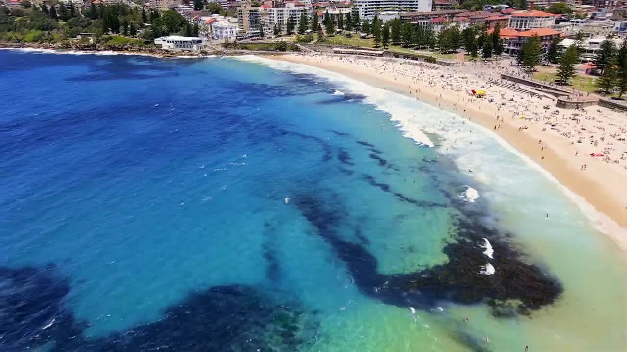 Waves break on the shore, where people are scattered across the sand. Dark patches of what looks like seaweed drift in the clear, turquoise shallows.
