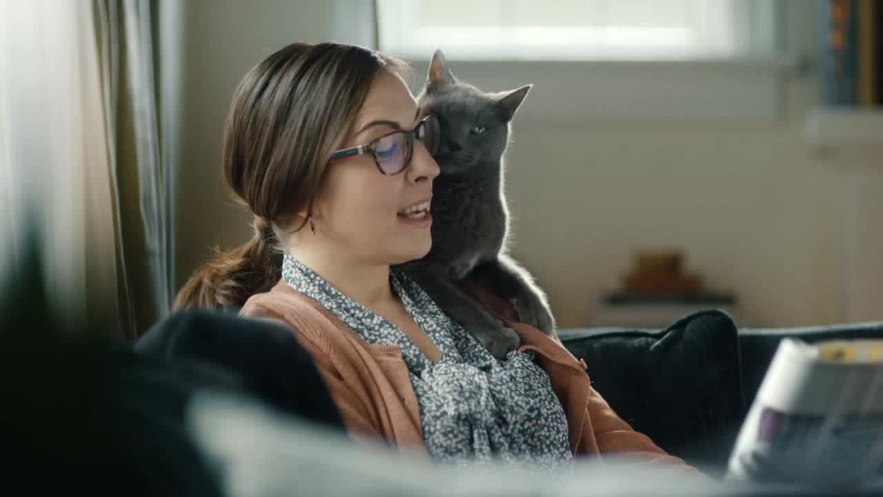 A woman with glasses is talking, a gray cat perched on her shoulder. The cat's gaze is fixed, its ears perked, as if listening intently.

