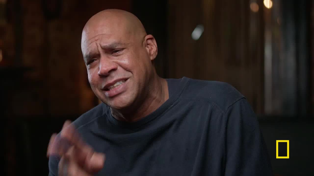 The man's hand gestures, as he speaks, are animated. His expression is a mix of seriousness and a hint of a smile.
