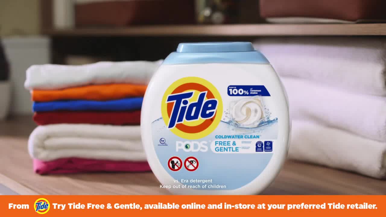 A bottle of Tide Pods sits front and center, with a stack of folded, brightly colored clothes to the left. Behind it, a neat pile of white towels rests on a shelf.
