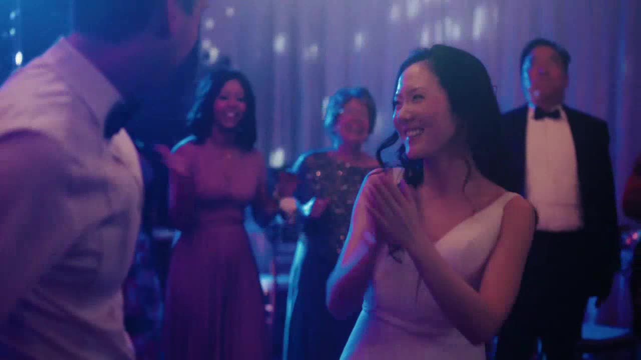 A woman in a white dress claps, smiling at someone out of view. Behind her, others in formal wear dance and celebrate.
