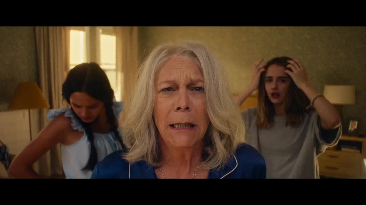 An older woman stares directly ahead, her face contorted in fear, while two younger women behind her clutch their heads, looking equally alarmed. The scene is set in a well-lit bedroom, likely in the United States, where a sense of dread hangs heavy in the air.
