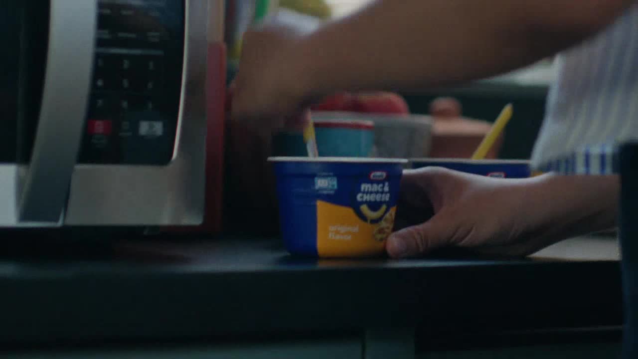A hand holds a blue and yellow cup of mac and cheese, while another hand stirs the contents with a yellow spoon. A microwave is visible in the background.
