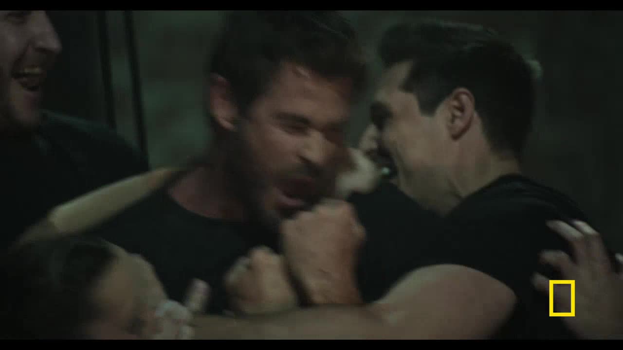 Two men are locked in a close embrace, their faces contorted in what looks like a struggle. A third person is partially visible, caught in the middle of the action.
