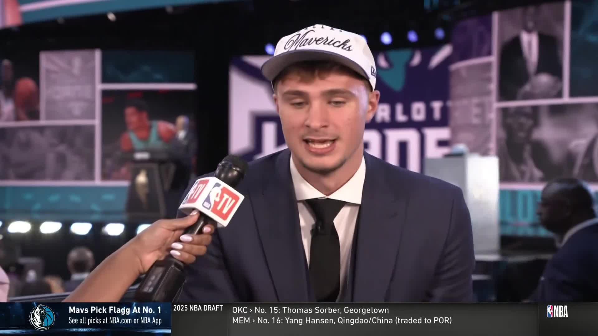 A young man in a suit and a Mavericks hat speaks into an NBA TV microphone. The background displays the Charlotte Hornets logo and images of past players.
