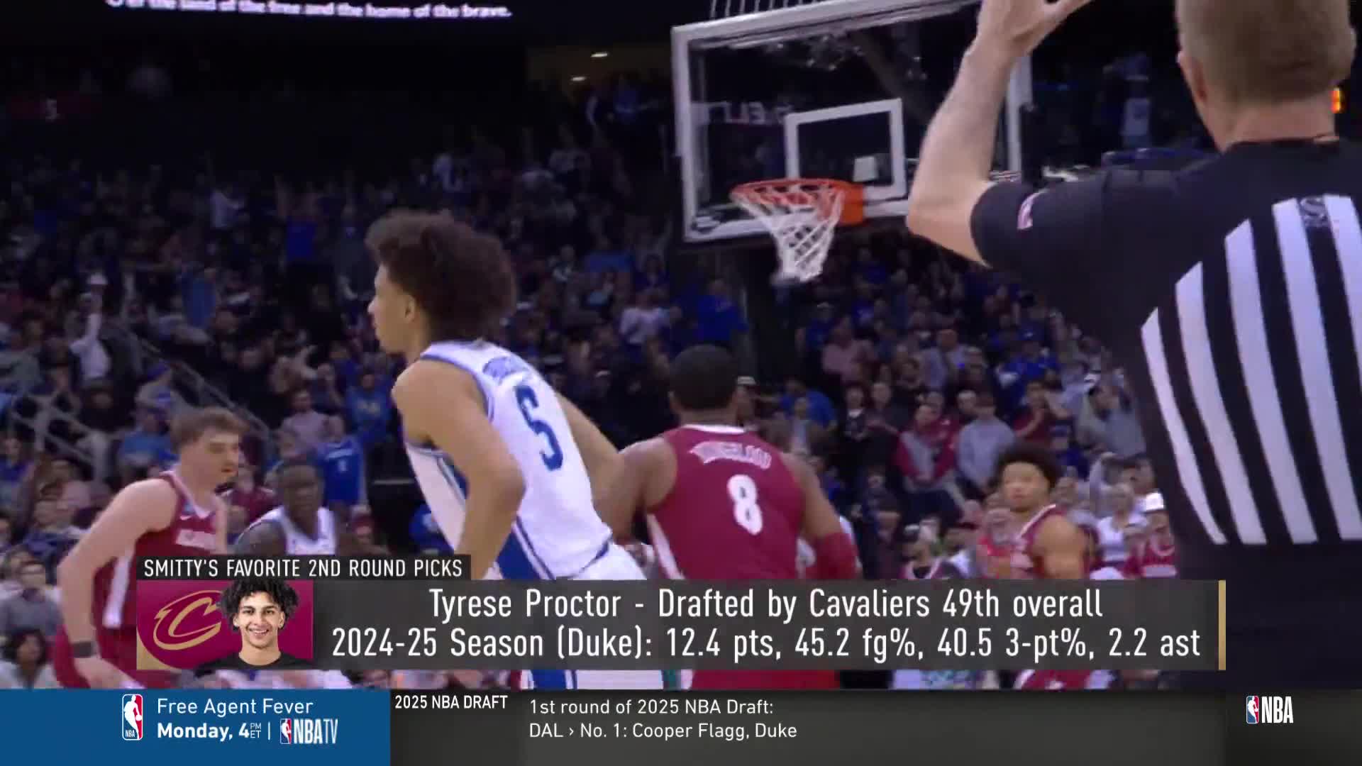 A player in a blue jersey with the number 5 looks toward the basket as the referee raises his arm. Another player in a maroon jersey is close behind him.
