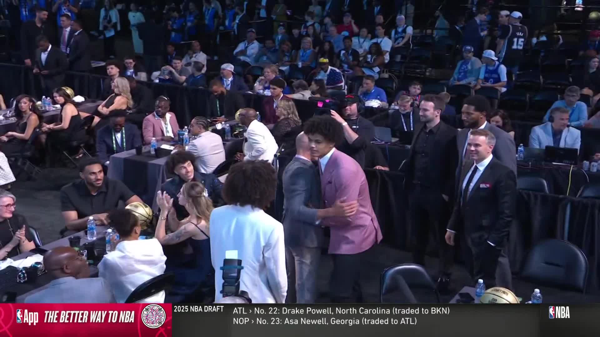 A young man in a pink blazer is embraced by someone in a gray suit, while others in the audience applaud. The scene unfolds during the NBA draft, as shown on NBA TV.
