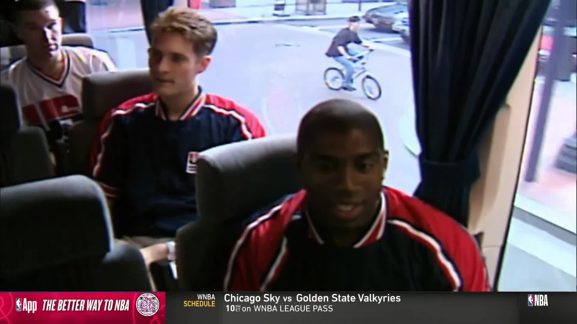 Three men in team jackets sit on a bus, while outside, a person on a bike rides down the street. The NBA TV graphic at the bottom indicates a WNBA game is about to start.

