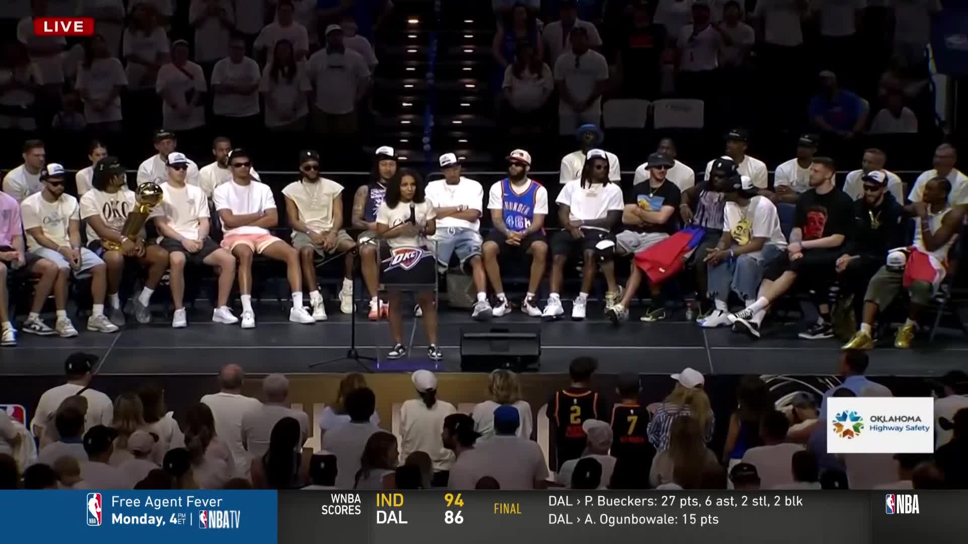A woman stands at a podium, speaking to a crowd while a group of men in casual clothes sit behind her. The scene is being broadcast on NBA TV, with the Oklahoma Highway Safety logo visible.
