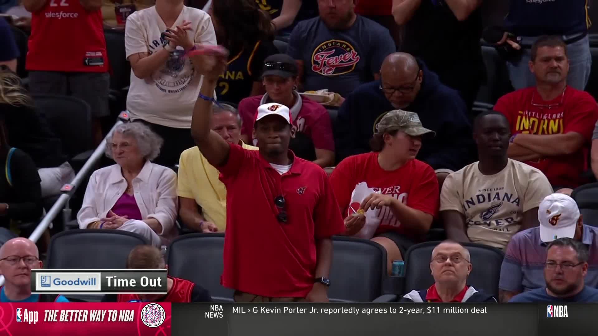 A fan in a red shirt and cap is holding up a small, pink item during a timeout. The crowd around him is watching, some clapping, while the NBA TV graphic displays the "Goodwill Time Out" logo.
