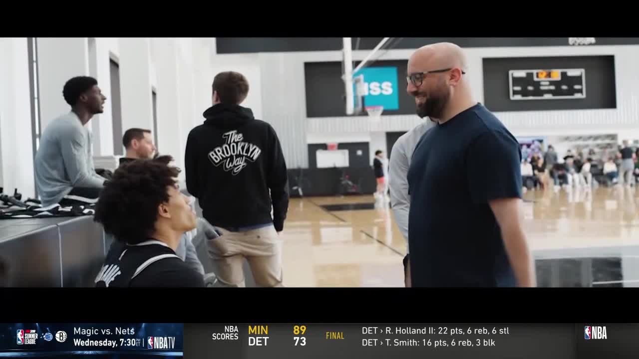 A coach in glasses smiles and chats with a player wearing a jersey. Other players are in the background, some sitting and others standing, all in a basketball court.
