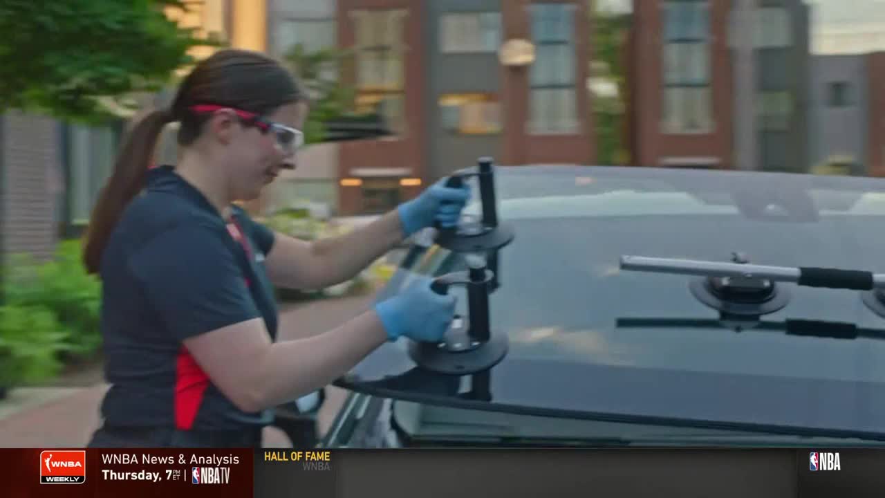 A woman in safety glasses and gloves carefully positions suction cups on a car's windshield. The NBA TV logo is visible in the corner of the screen.
