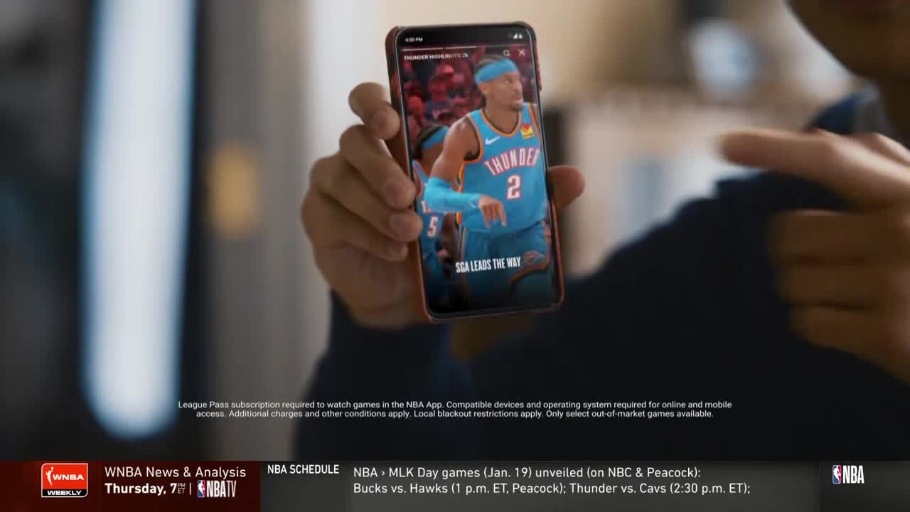 A hand holds a phone displaying an NBA game, the screen showing a player in a teal jersey. The bottom of the screen reads "SGA LEADS THE WAY".
