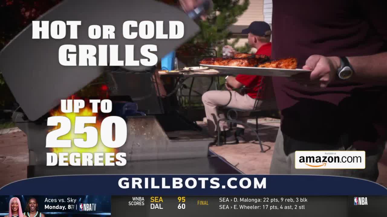 A man carries a plate of grilled food towards someone sitting at a table. The scene is a backyard cookout, with a grill open and ready.
