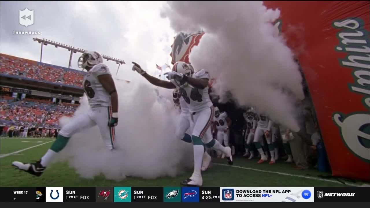Dolphins players burst from the tunnel into a cloud of smoke, their white and teal uniforms standing out. The players, energized, point and gesture as they head onto the field.
