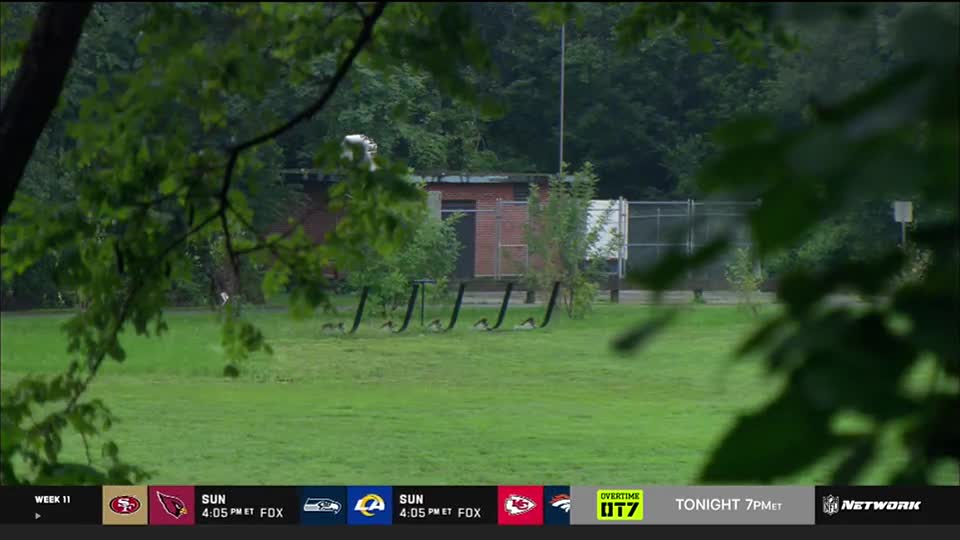The camera is positioned behind some foliage, looking across a grassy field towards a small brick building and a fence. The NFL Network logo and game information are displayed at the bottom of the screen.
