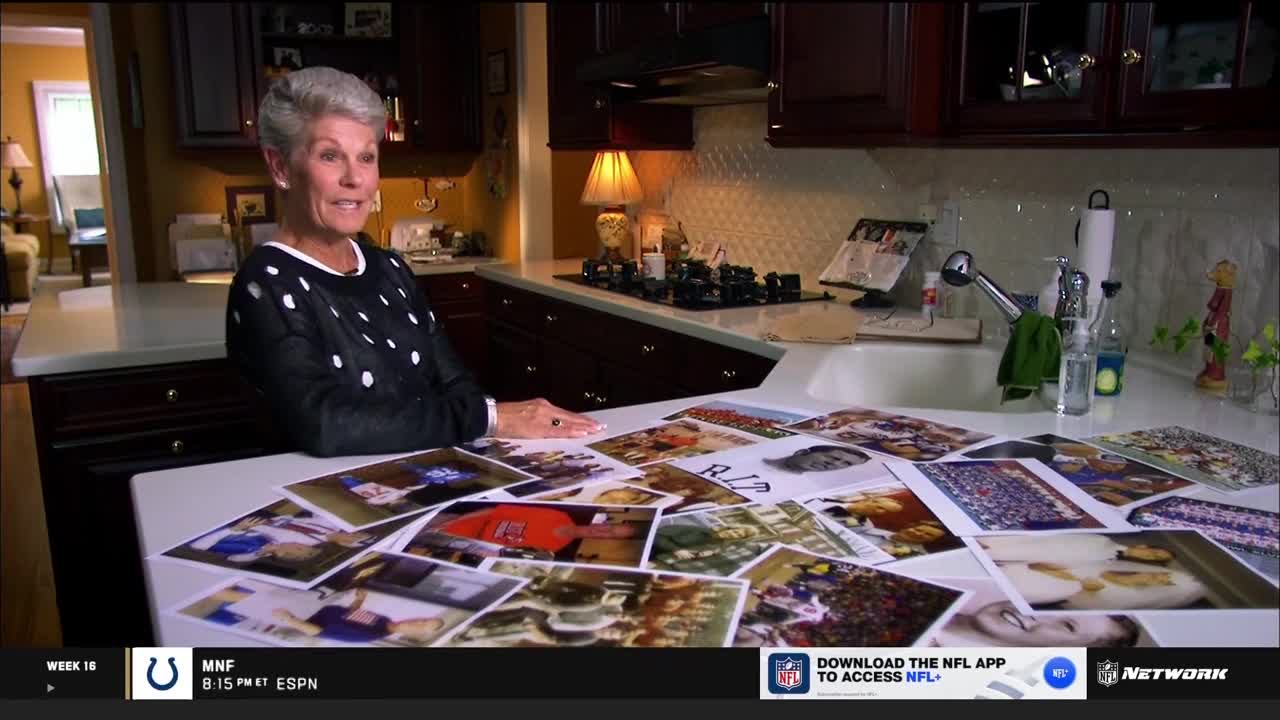 A woman in a polka-dot sweater sits at a kitchen counter, smiling as she looks towards the camera. Scattered across the counter are numerous photos, some of which appear to be related to football.
