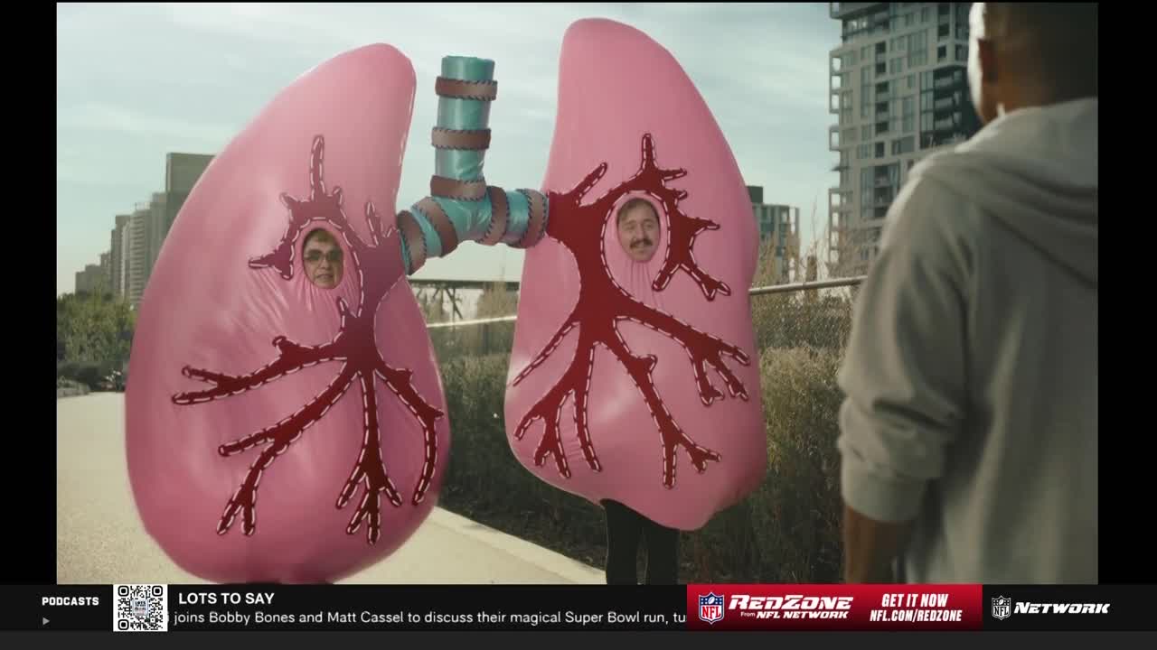 Two people in pink lung costumes stand outside, connected by a turquoise tube. A man in a gray hoodie looks on as the "lungs" face the camera.
