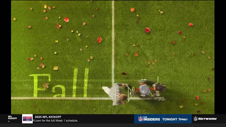 A groundskeeper, atop a small machine, is mowing the field, with the word "Fall" painted on the grass. Scattered leaves, in shades of red and yellow, are scattered across the green.
