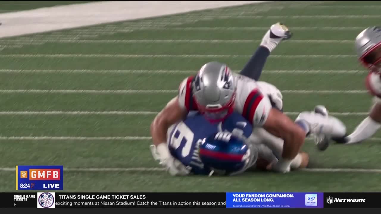 A player in a Patriots uniform has wrapped up a Giants player, driving him to the ground. The Giants player's helmet is turned, and the Patriots player is on top, his helmet visible.
