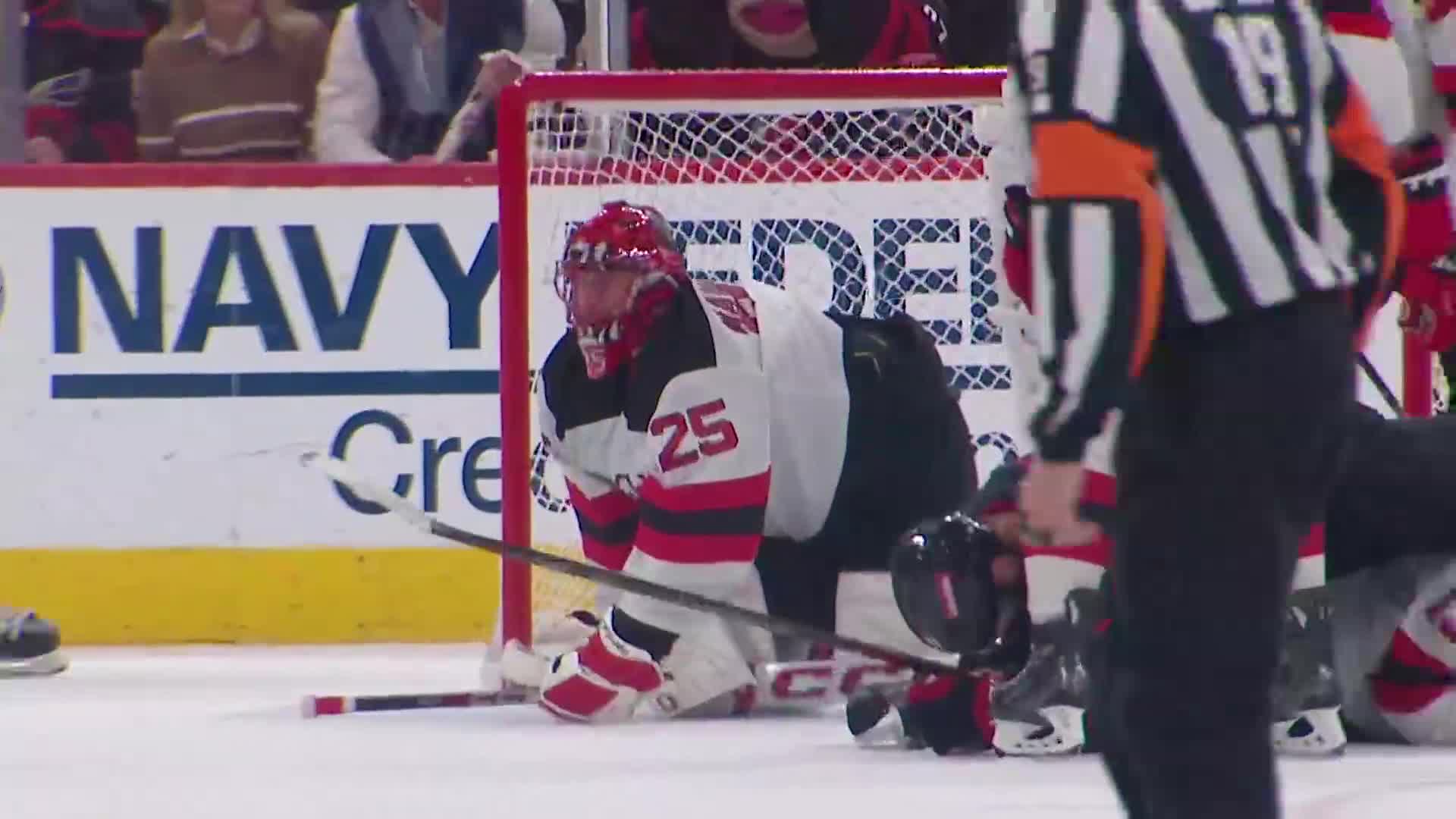 The goalie, in a red helmet and jersey, is down on his knees in front of the net. A player is on the ice nearby, and the referee is standing over them.
