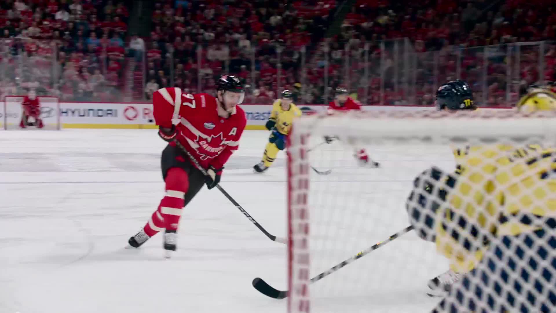The player in red skates toward the net, stick extended, while a player in yellow pursues. A fallen player in red lies on the ice near the goal.

