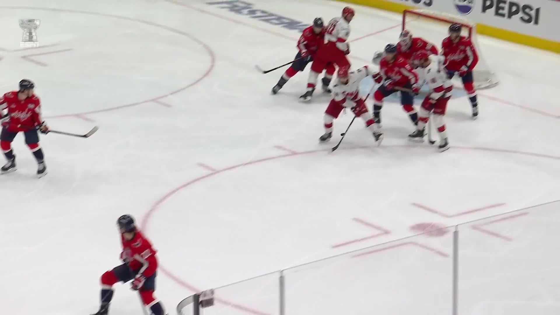 A scrum of red-clad players swarms the net, battling for position. Two players in blue and white are skating in from the left, trying to join the fray.
