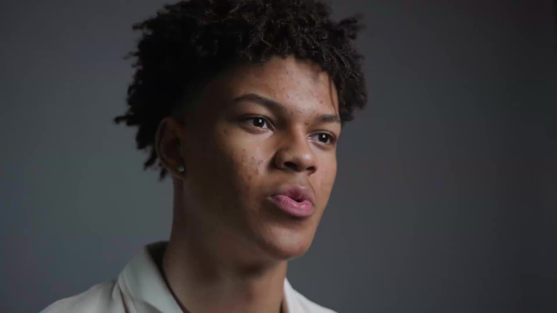 A young man with dark, curly hair is speaking, his lips slightly parted. He's facing the camera, and the background is a muted gray.
