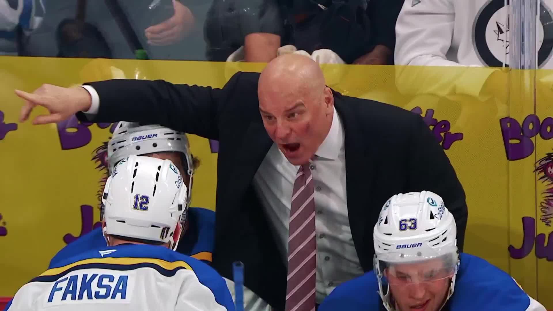 The coach is yelling, his arm outstretched, pointing toward the ice. The players in blue jerseys listen intently, their helmets and faces visible.
