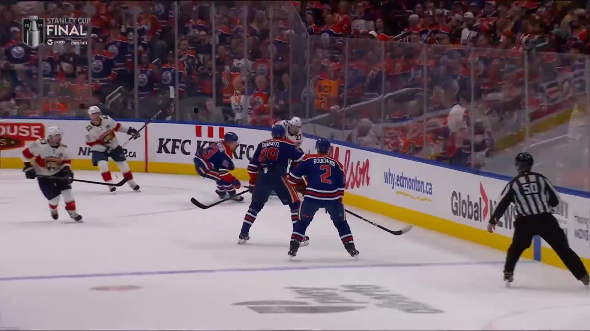 Two players in blue jerseys are battling near the boards, while a player in a white and red jersey skates toward them. The referee, in black and white stripes, has his arms out, watching closely.
