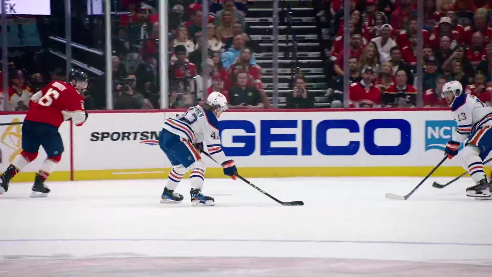 The player in blue and white skates toward the puck, stick low and ready. A player in red and black is closing in fast, trying to catch up.
