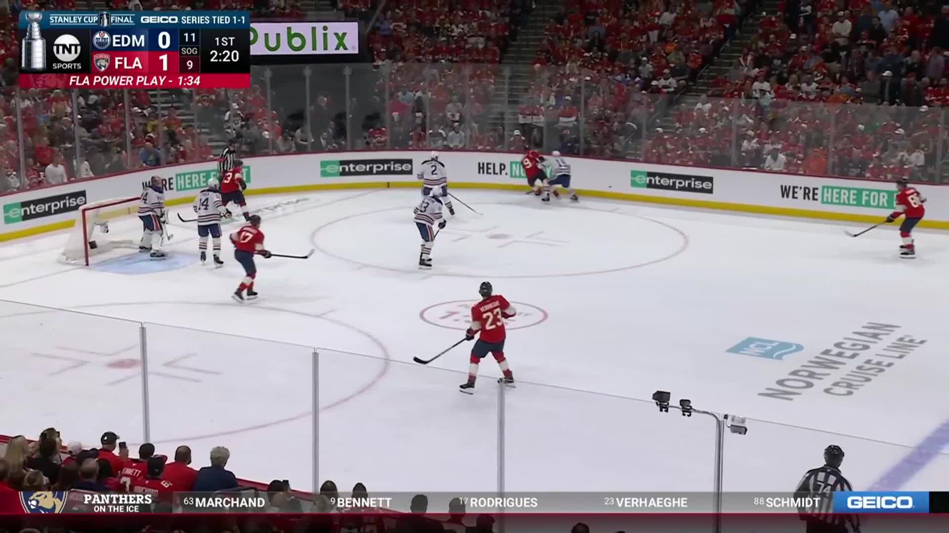 A Florida Panthers player in red skates toward the puck, stick raised. The Edmonton Oilers are in blue and white, defending in their zone during a power play.
