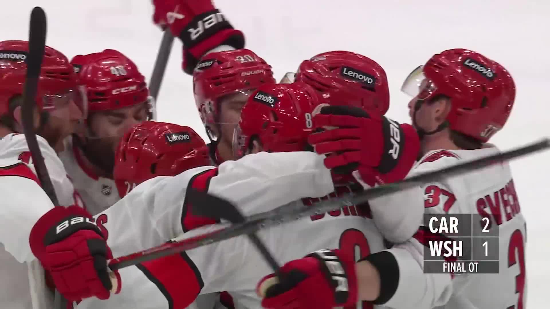 The Carolina Hurricanes players are huddled together, their red helmets and gloves a bright contrast to their white jerseys. Sticks are held aloft as they celebrate a hard-fought overtime win.
