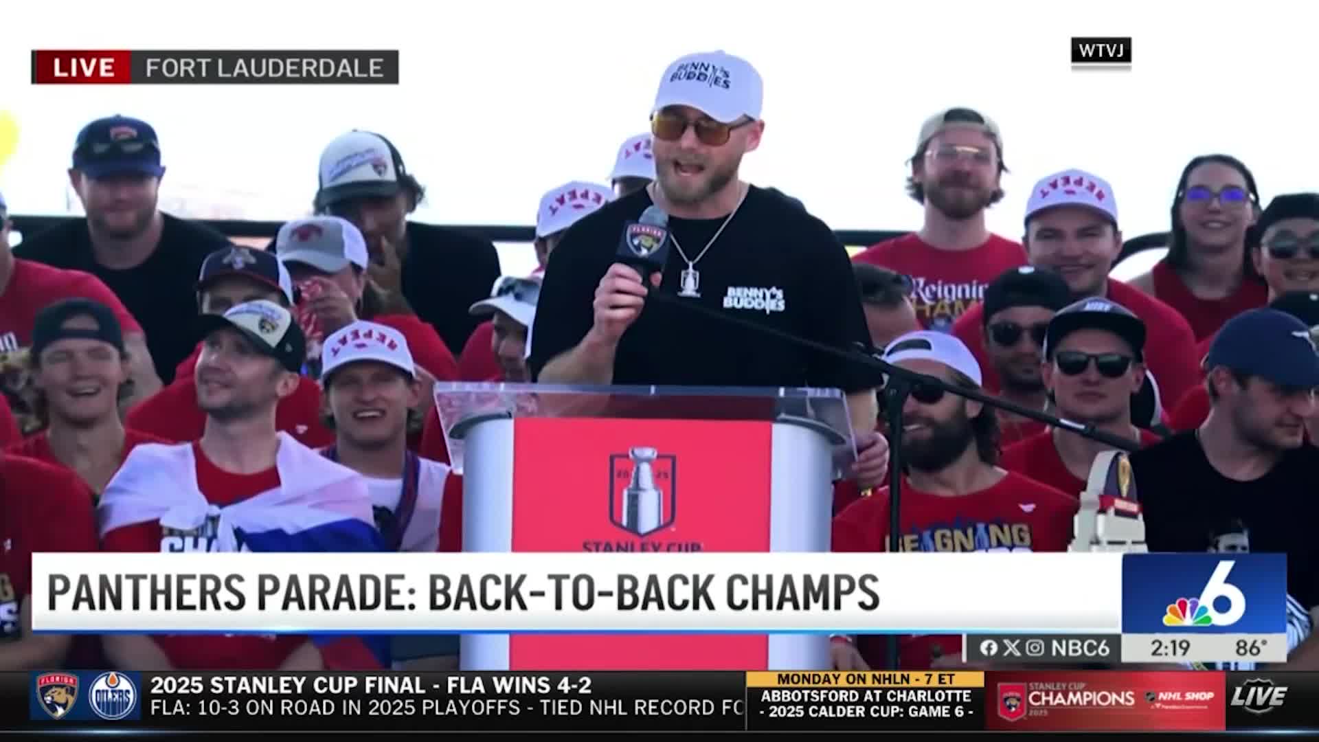 A man in sunglasses and a white hat speaks into a microphone from a podium, with a crowd of cheering people behind him. The podium displays the Stanley Cup logo, and the broadcast is live from Fort Lauderdale.
