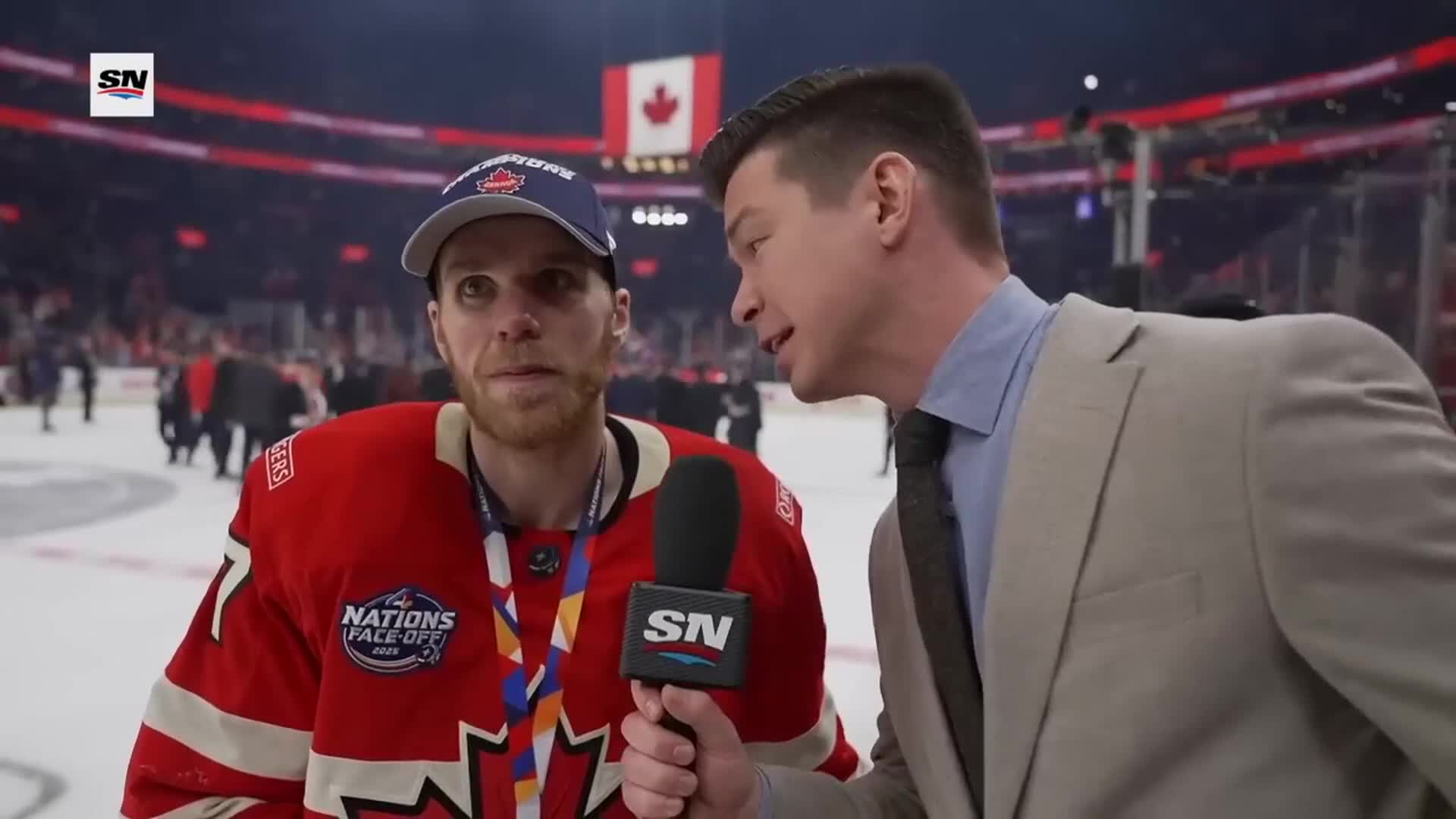 A reporter in a suit holds a microphone toward a hockey player wearing a red jersey and a championship hat. The player, still wearing a medal, looks directly at the camera, his expression a mix of exhaustion and pride.
