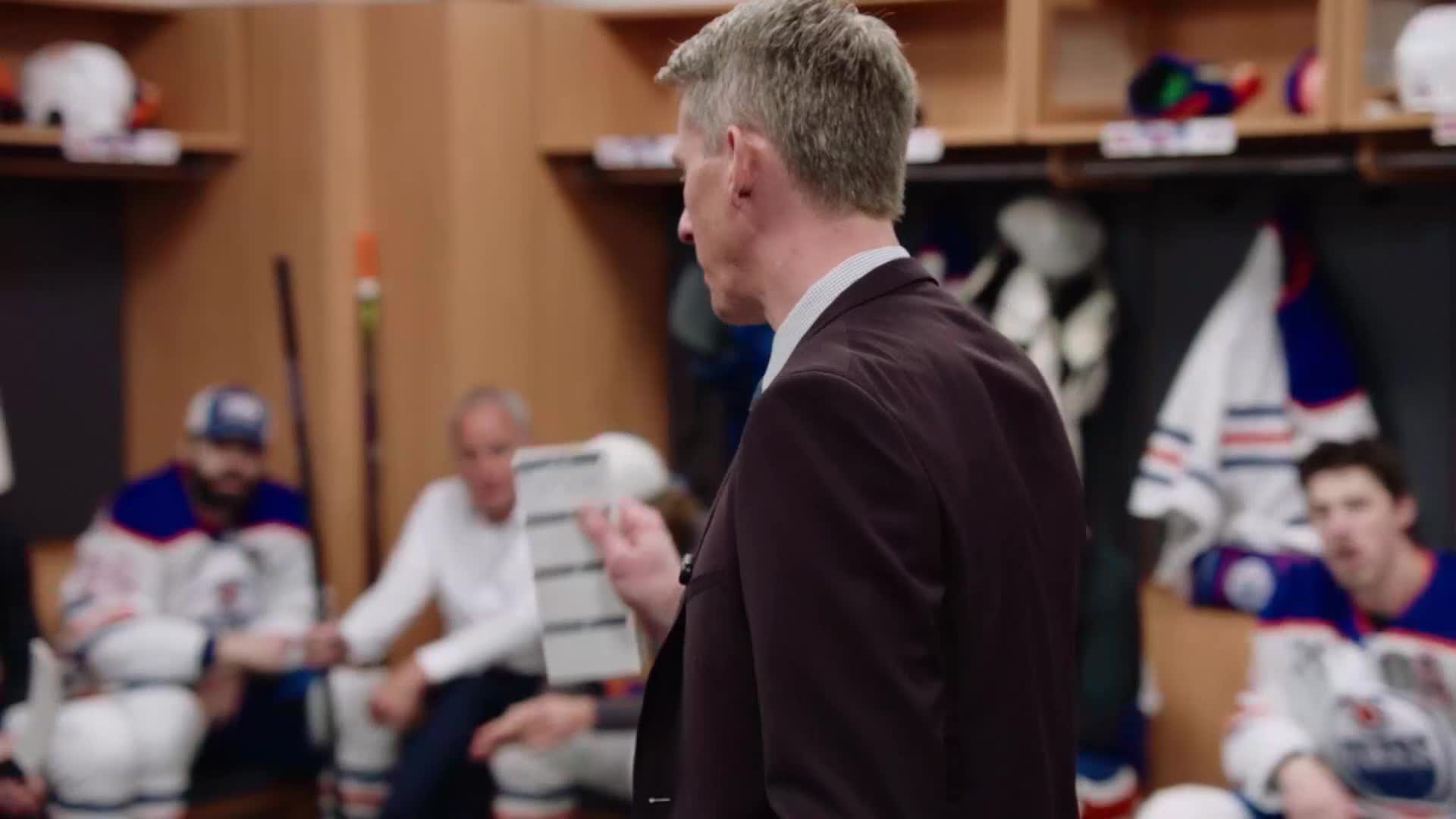 A man in a suit reads from a sheet of paper, addressing a group of hockey players in their locker room. The team, dressed in blue and white jerseys, listens intently.
