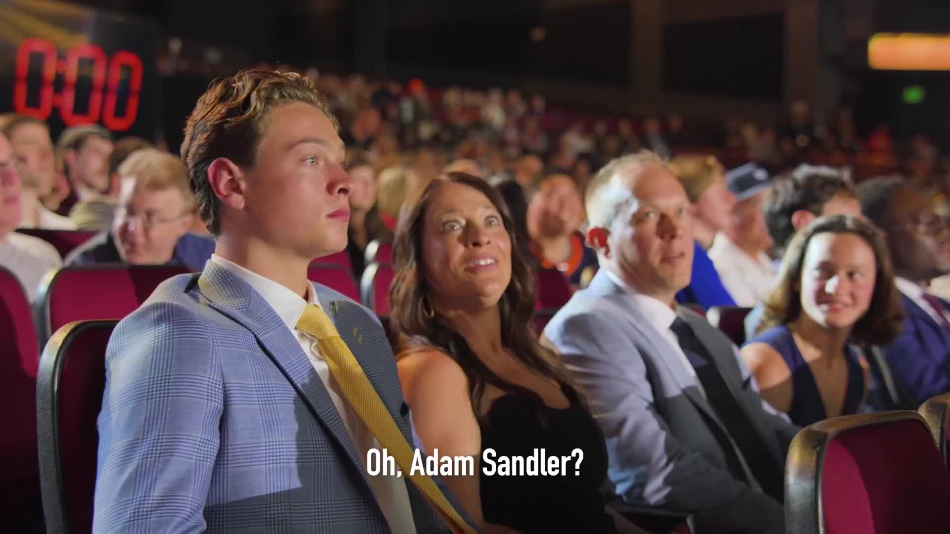 A young man in a light blue suit and yellow tie stares intently at something off-screen. Beside him, a woman's face lights up with surprise as she looks in the same direction.
