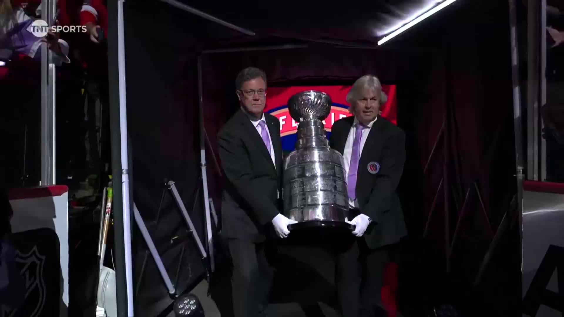 Two men in suits and white gloves carefully carry the Stanley Cup from a dark entrance. The iconic trophy gleams under the bright lights.
