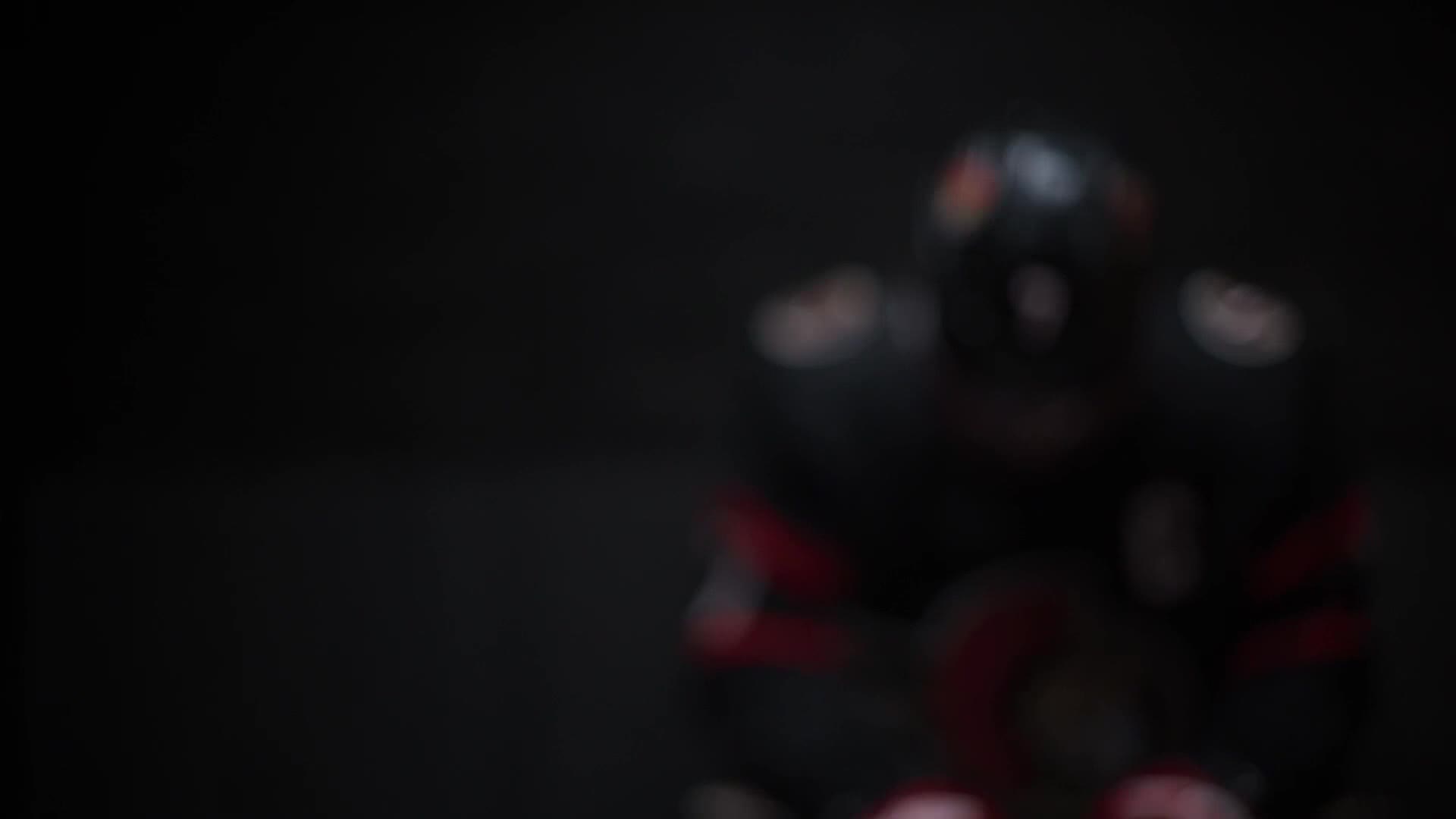 A player in black and red gear is skating toward the camera, a dark silhouette against the rink. The background is a deep, inky black.
