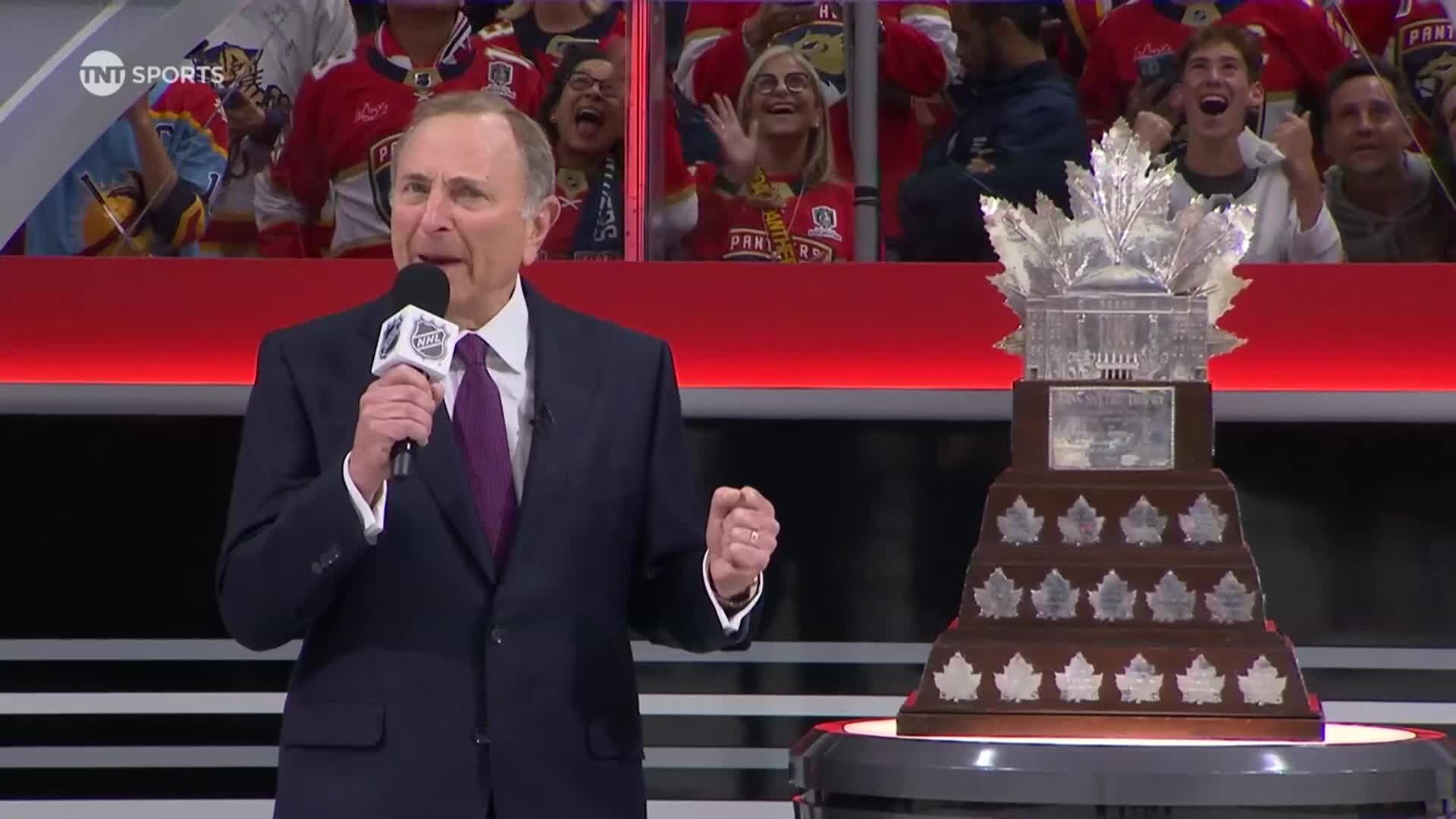 The man in the suit holds a microphone, addressing a crowd, with the Stanley Cup gleaming on a pedestal to his right. Cheering fans in the stands behind him are visible.
