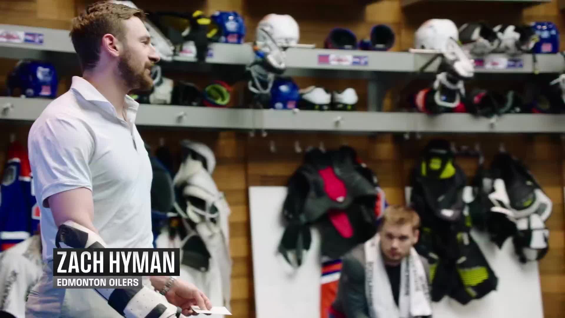 Zach Hyman, in a white polo, stands in the locker room holding a card, while another player sits in the background. Hockey gear, including helmets and pads, lines the shelves behind them.
