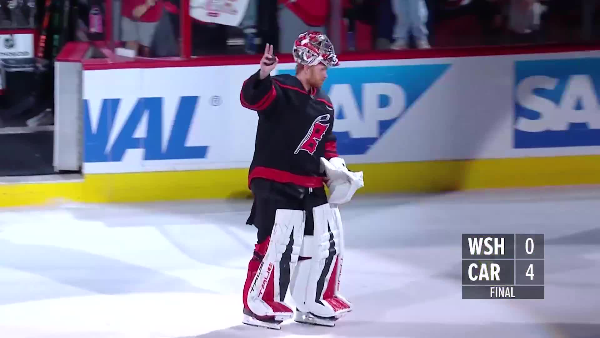 The Carolina Hurricanes goalie, still in full gear, skates toward the center of the ice, giving a peace sign to the crowd. The scoreboard shows a shutout win for the Canes on NHL Network.
