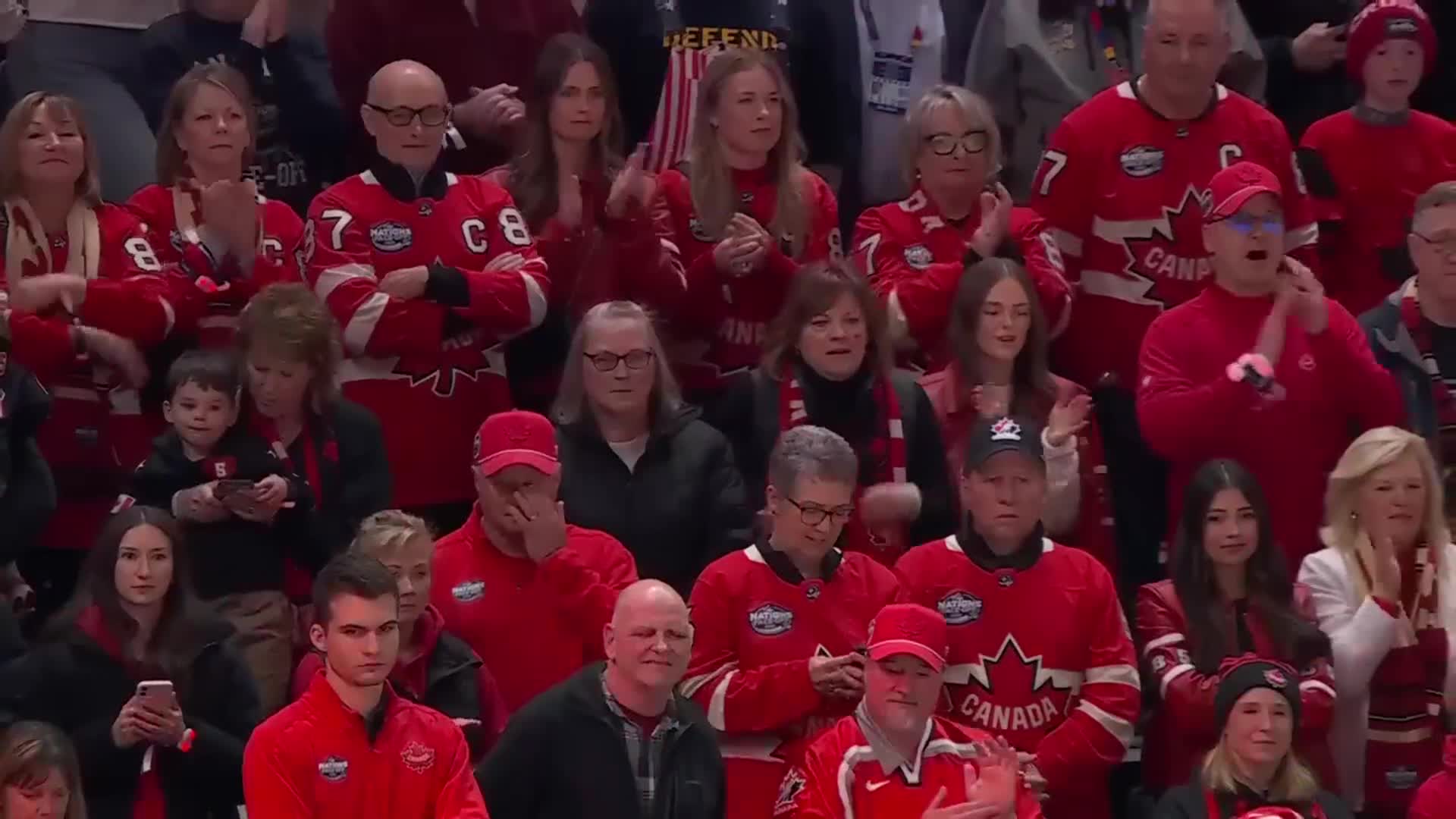 A crowd of people in red jerseys and hats are clapping and cheering, some with their hands raised. Several individuals appear to be watching a hockey game, some with expressions of excitement or anticipation.
