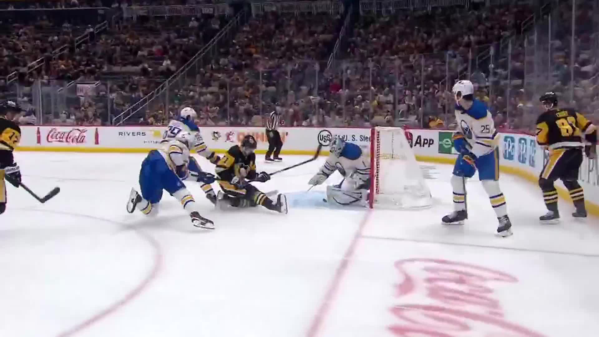 A player in blue and white skates toward the net, while a player in black and gold is down on the ice. The goalie is in position, ready to block the puck.
