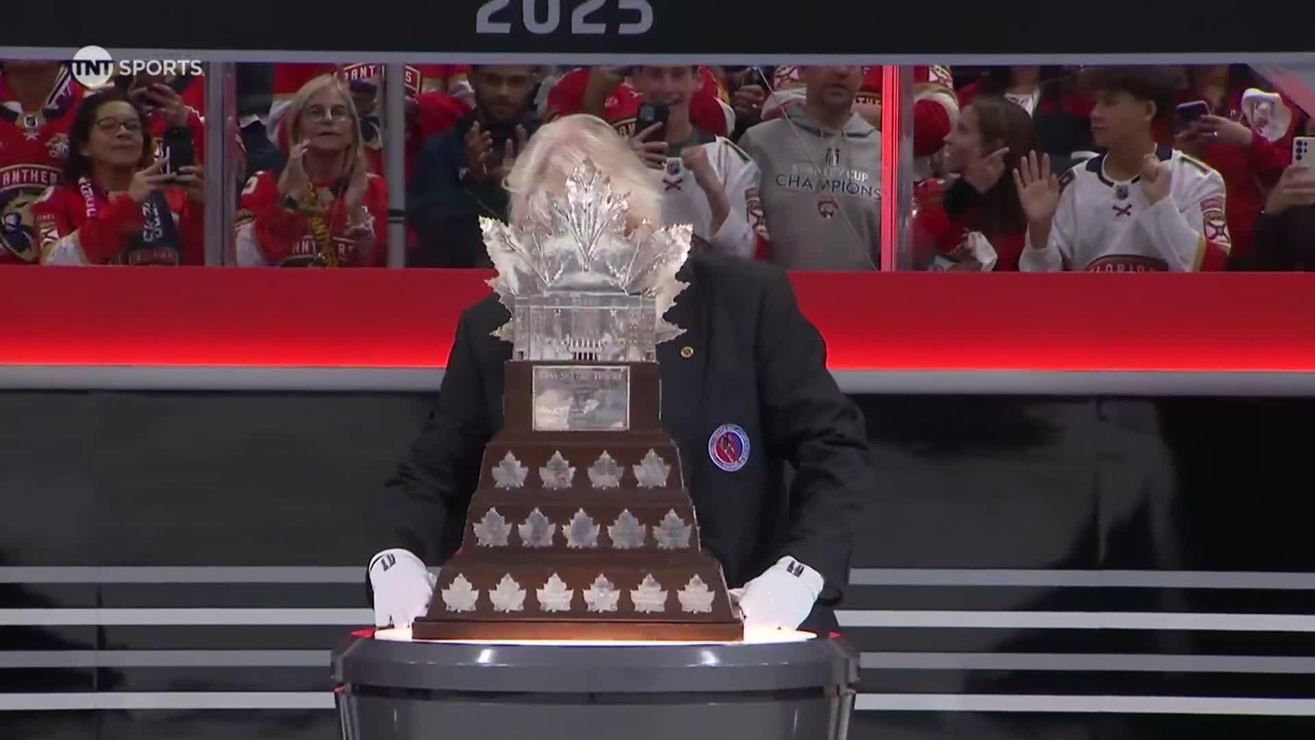 A man in a suit, wearing white gloves, is carefully holding the Stanley Cup. The crowd in the background is cheering, many with their phones up.
