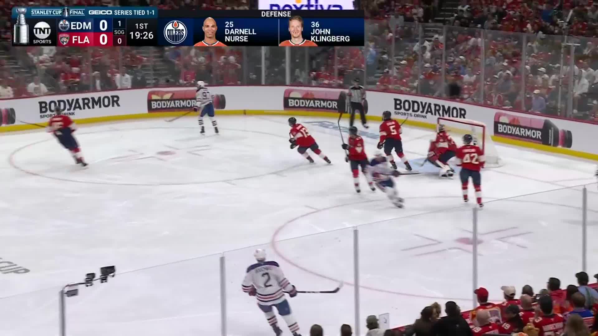 The Oilers player in blue skates toward the net, pursued by a cluster of red-clad Panthers. The puck is in the air, headed toward the goal.
