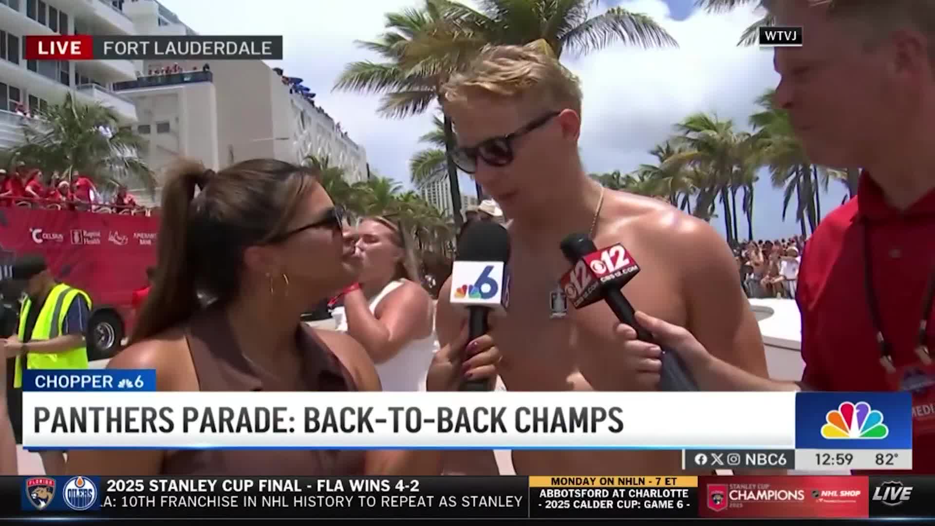 A shirtless man with sunglasses speaks into two microphones, while two women listen intently. Behind them, a parade float with red decorations and people is visible, and palm trees sway in the background.
