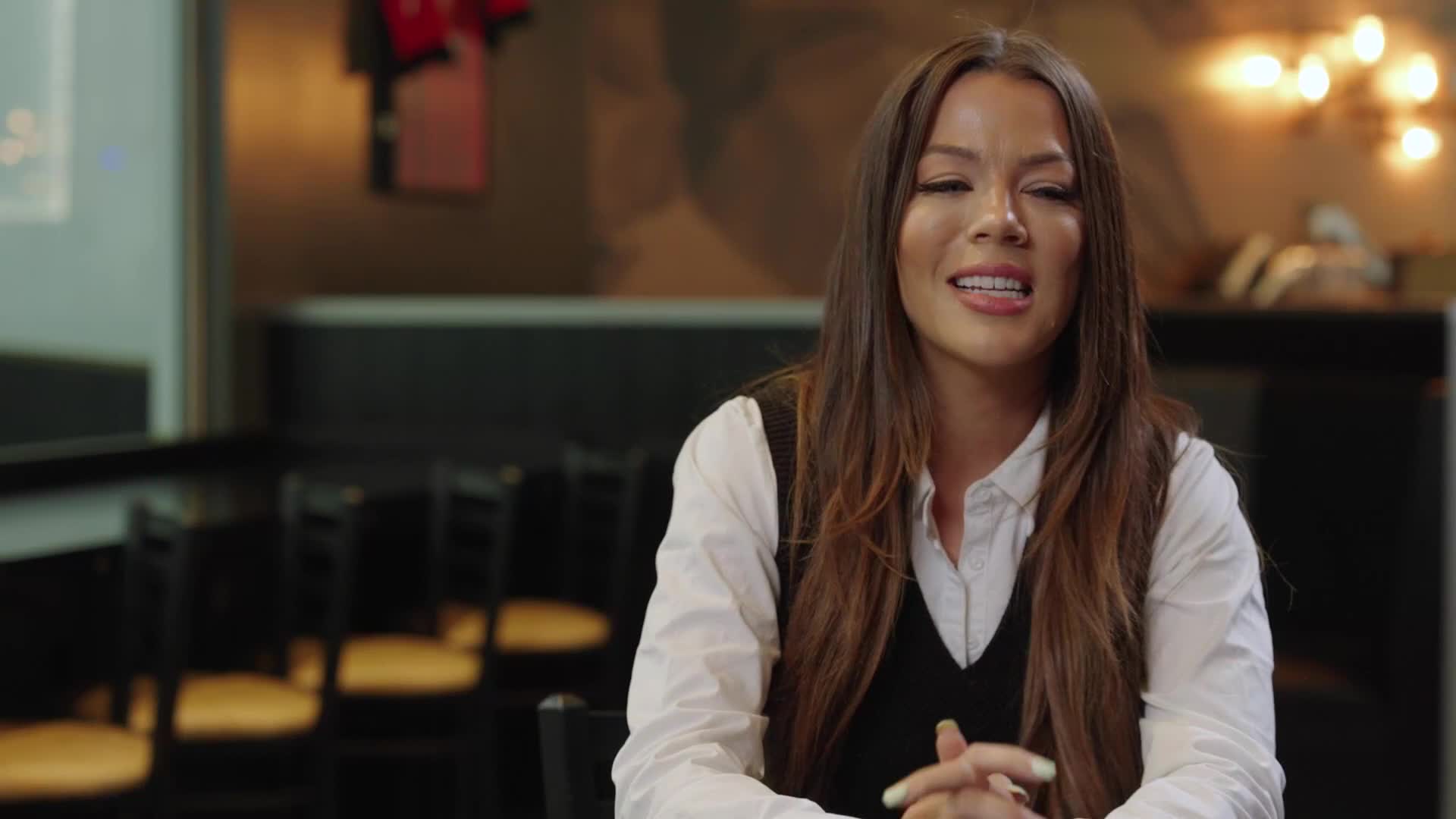She's seated in a restaurant, hands clasped in her lap, speaking directly to the camera. The lighting is warm, and behind her, the space is softly out of focus.
