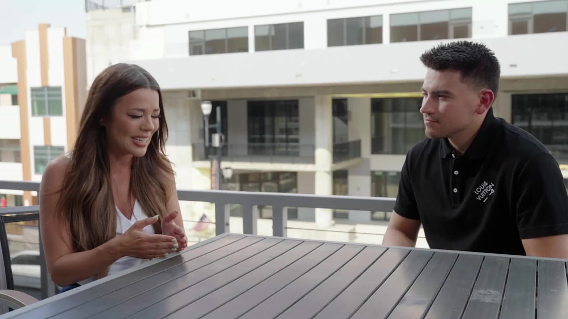 On a sunny patio, a woman gestures with her hands as she speaks, while a man in a black polo shirt listens intently. They're seated at a dark table, with a modern building visible in the background.
