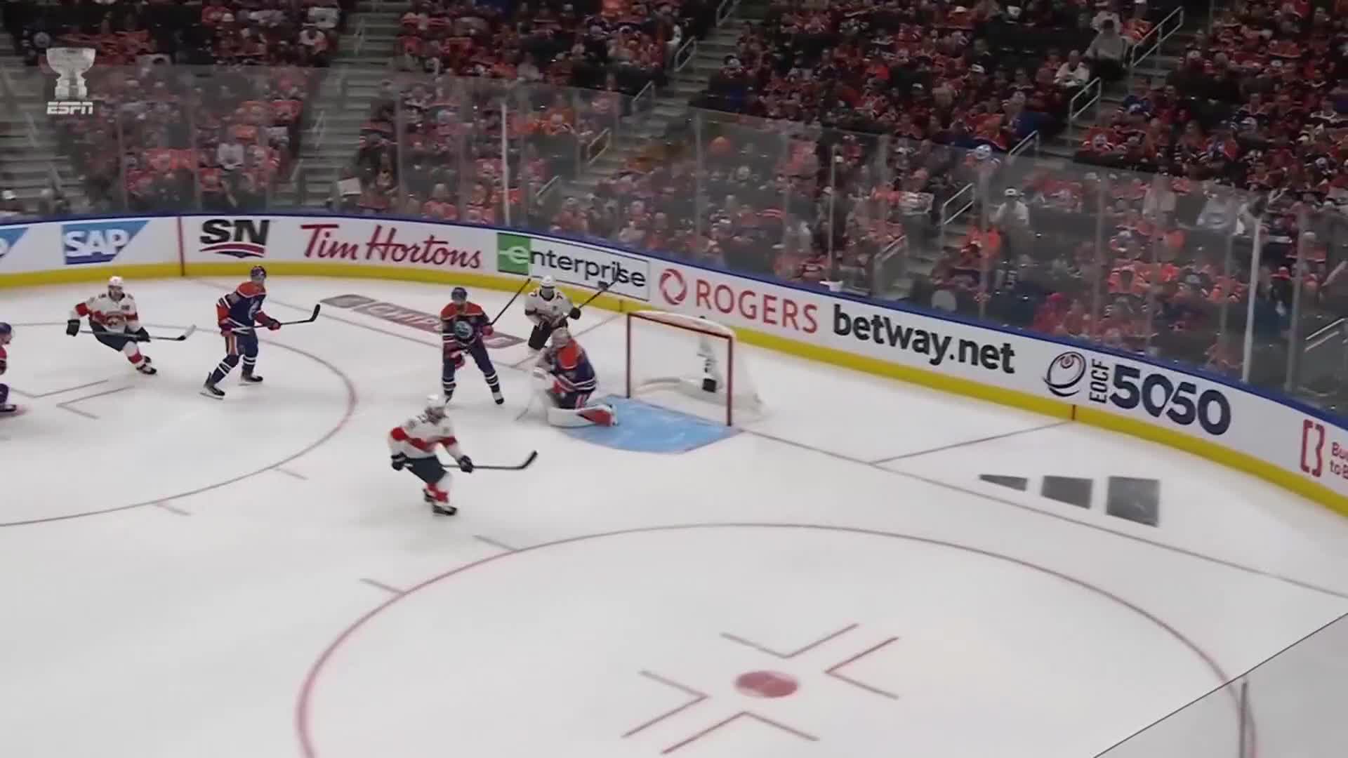 The goalie is down, guarding the net as a player in a red and white jersey skates toward him. Several players in blue and orange jerseys are positioned near the center of the ice.

